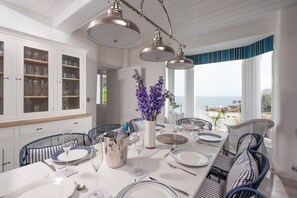 Ein Essbereich mit einem langen Tisch, gedeckt für vier Personen, umgeben von Korbstühlen, einer weißen Kommode mit Glastüren und Blick auf das Meer.