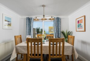 A dining area with a table set for four, wooden chairs, a chandelier, and a view of the sea through large windows. - 1 Golden Bay is a stylish ground floor apartment offering sea glimpses (Newquay)