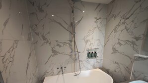 A modern bathroom with a freestanding bathtub, a glass shelf with toiletries, and marble-patterned walls. - Luxury 4-Bedroom Apartment near Oxford St/ Baker Street (London)