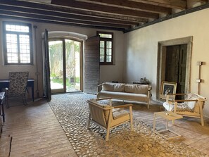 Una habitación rústica con suelo de piedra, un sofá, una silla y una mesa. Hay dos ventanas y una puerta que da al exterior.