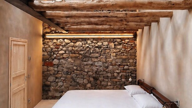 Une chambre avec un mur en pierre, des poutres en bois, un lit et une lucarne.