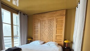 Een slaapkamer met een groot bed, een houten hoofdbord met ingewikkelde houtsnijwerken, een nachtkastje met een lamp en een raam met gordijnen.