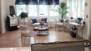 Ein modernes Wohnzimmer mit einer Couch, Sesseln, einem Couchtisch und einer TV-Konsole. Es gibt Fenster mit Vorhängen, eine Zimmerpflanze und einen kleinen Tisch mit Gegenständen darauf.
