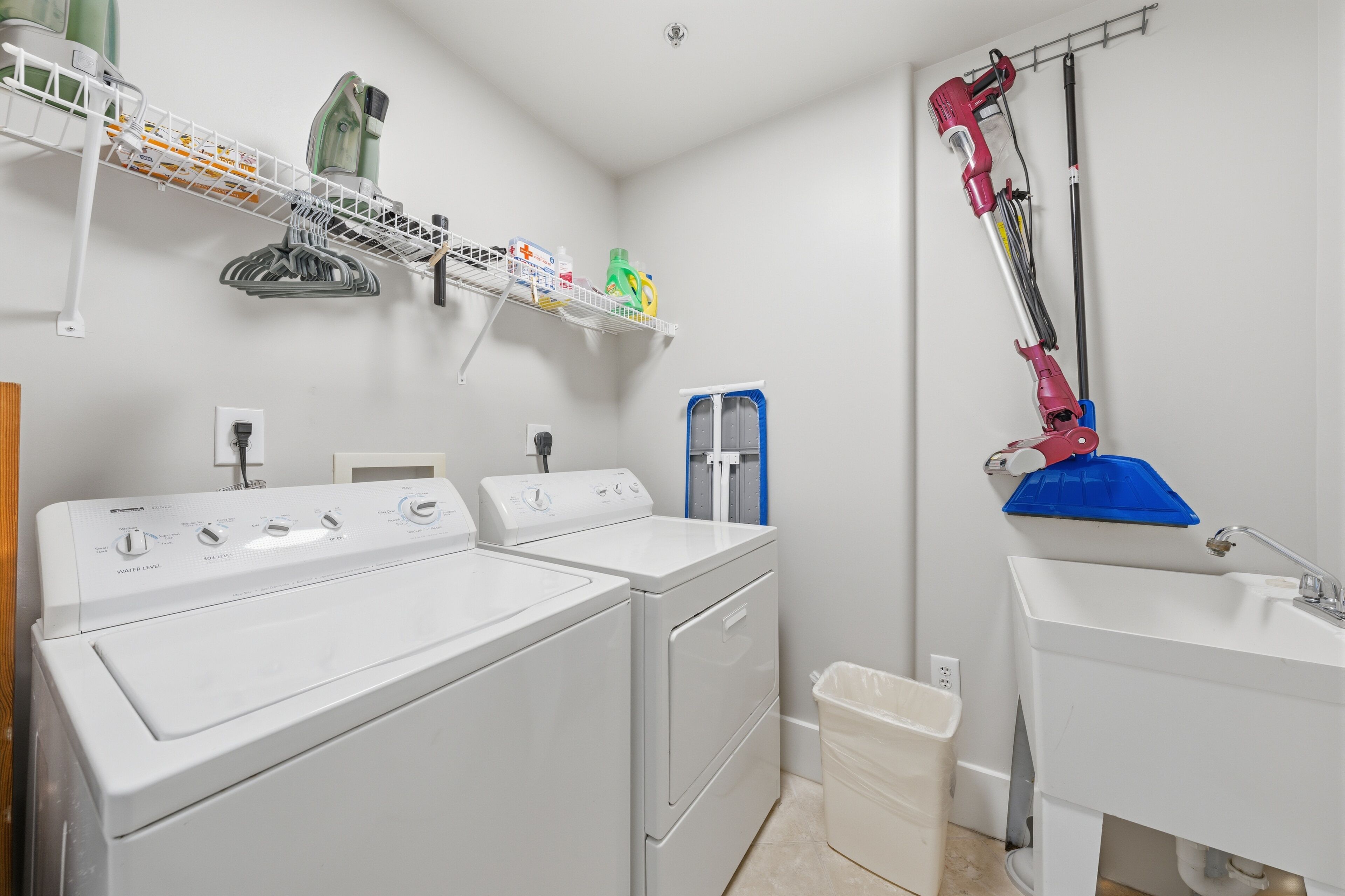 A laundry area with two washers and dryers, a shelf with cleaning supplies, a blue bucket, and a sink.