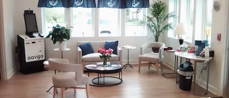 A modern living room with a sofa, armchairs, a coffee table, and a TV stand. There are windows with curtains, a potted plant, and a small table with items on it.