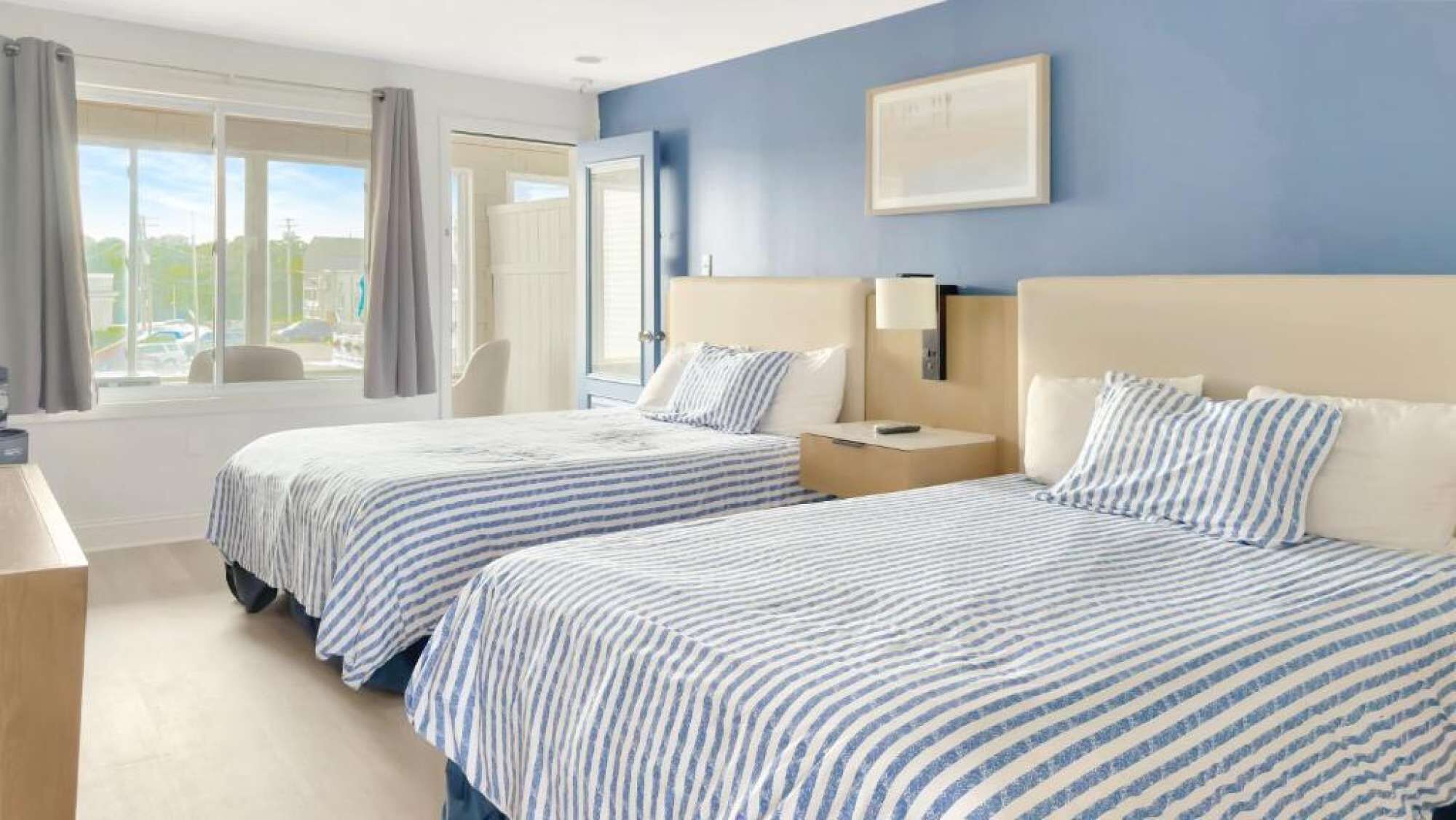 Une chambre d’hôtel avec deux lits, un bureau, une chaise, une fenêtre avec des rideaux et un tableau encadré au mur.
