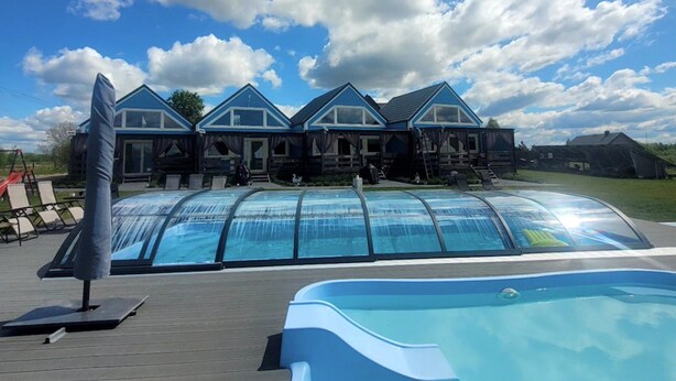 Una piscina con cerramiento de vidrio, una terraza de madera y un edificio con múltiples ventanas y tejado azul.