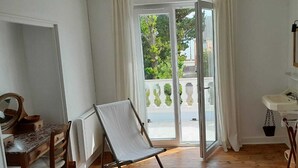 Ein Zimmer mit Holzboden, einer Holzkommode, einem weißen Waschbecken und einem Balkon mit Blick auf Grünflächen und ein Gebäude.