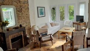 Ein gemütliches Wohnzimmer mit Kamin, weißen Möbeln, einem Fernseher und großen Fenstern mit Blick auf Grünflächen.