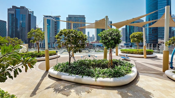 Une place urbaine moderne, agrémentée d’un aménagement paysager géométrique comprenant des jardinières et des arbres, et entourée d’immeubles de grande hauteur.