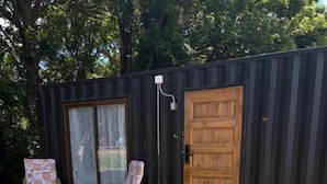 Una terraza de madera que conduce a una estructura de metal negro con una puerta de madera, dos sillas estampadas y un pequeño jardín con rocas.