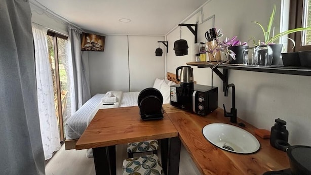 Una cocina compacta con una mesa de madera, un lavabo blanco, una cafetera y una estantería con utensilios de cocina y plantas.