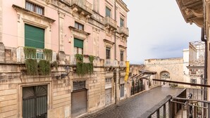 Eine enge Straße mit einem Gebäude, das einen heruntergelassenen Rollladen, einen Balkon mit Pflanzen und einem Metallgeländer zeigt.