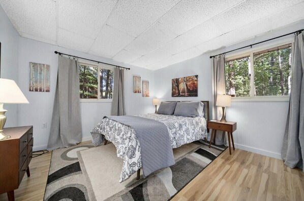 A bedroom with a bed, two bedside tables with lamps, a dresser, a window with curtains, and artwork on the walls.