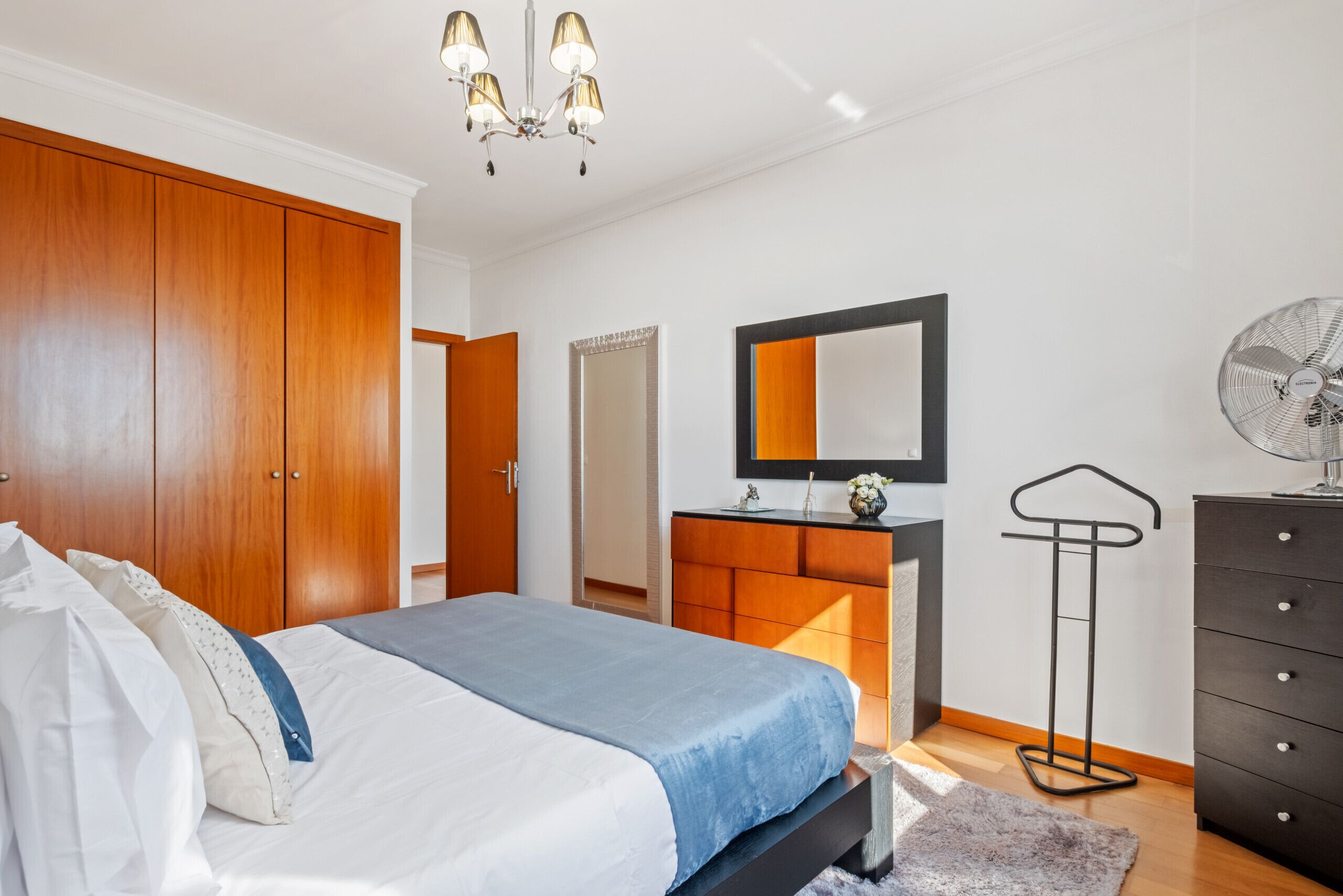 A bedroom with a bed, wooden wardrobe, dresser, mirror, and a standing fan.