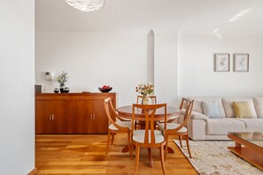 A modern living room with a round wooden dining table, wooden chairs, a beige sofa, and a white wall with two framed pictures. - Cani o Quintas V by Homie (Santa Cruz)
