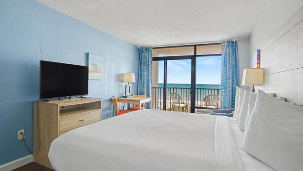 Habitación de hotel con una cama grande, un televisor sobre un mueble de madera, un escritorio con una silla y un balcón con vista al océano.