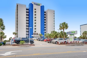 A multi-story hotel with a prominent 'OceanPark' sign, a parking lot with various vehicles, and palm trees. - Oceanfront | Kitchen | Dog Friendly | Sleeps 4 (Myrtle Beach)