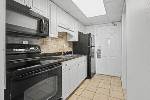 A compact kitchen with white cabinets, a black oven, a microwave, a refrigerator, and a tiled floor.