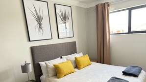 Un lit bien fait, recouvert d’une couvre-lit à motifs floraux, avec deux oreillers et deux serviettes pliées. Une petite lampe de chevet est posée sur une table de chevet à côté du lit. Deux reproductions botaniques encadrées sont accrochées au mur, au-dessus du lit.