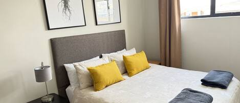 Una cama bien hecha con una colcha estampada, dos almohadas y dos toallas dobladas. Una lámpara de noche está sobre una mesita junto a la cama. Dos láminas botánicas enmarcadas cuelgan en la pared encima de la cama.