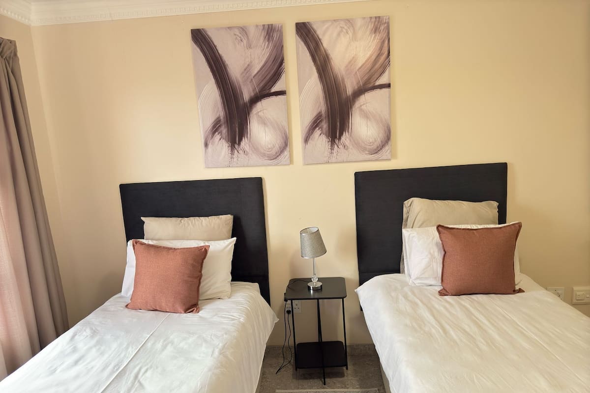 A hotel room with two beds, each with white bedding and brown pillows, a small table with a lamp, and abstract art on the wall.