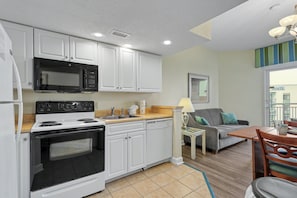 A compact kitchen with white cabinets, a microwave, stove, and dishwasher. There's a dining area with a table and chairs, and a sofa in the background. - HS-0823 8th Fl Ocean Retreat | Pools | Lazy River (Myrtle Beach)