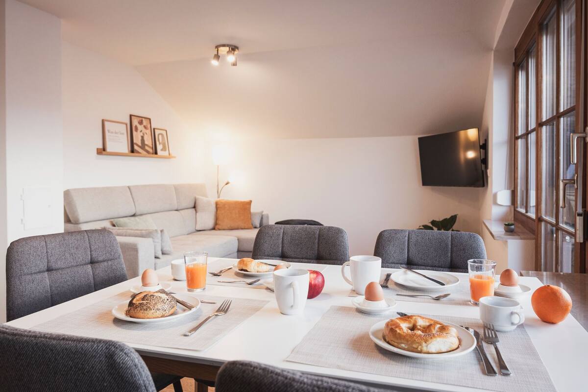 A dining area with a table set for breakfast, a sofa, and a TV mounted on the wall.