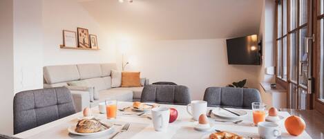 Spiseområde med et bord dækket til morgenmad, en sofa og et fjernsyn ophængt på væggen.