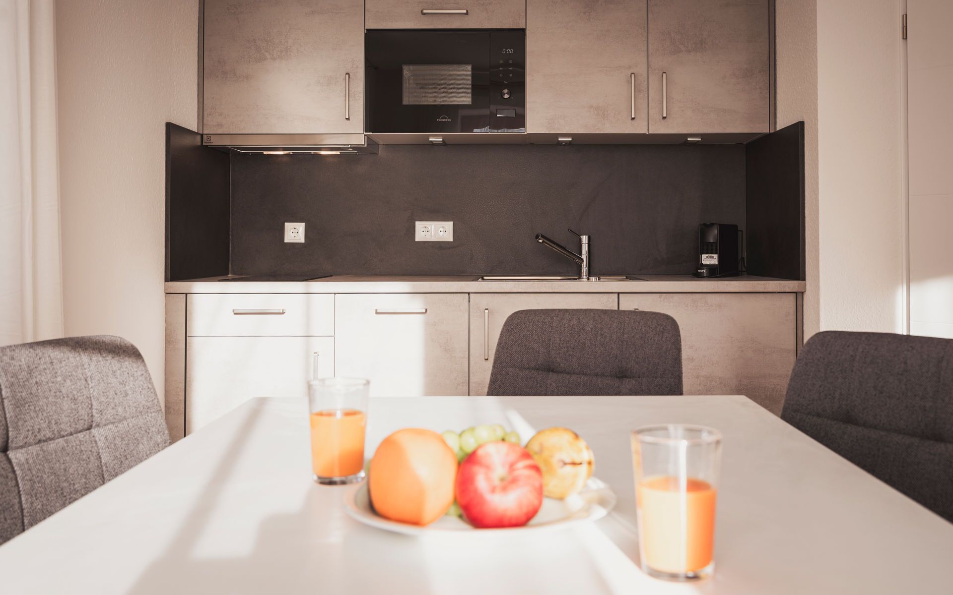 Eine moderne Küche mit Mikrowelle, Backofen und Kaffeemaschine. Ein Esstisch mit einer Fruchtschale und zwei Gläsern Saft.