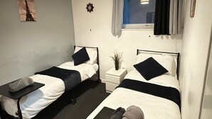 Ein Doppelzimmer mit zwei Betten, einem kleinen Tisch, einem Nachttisch, einer Wanduhr, einem Fenster mit Vorhängen und einer Deckenleuchte.