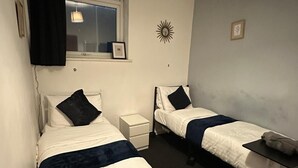 Ein Doppelzimmer mit zwei Betten, einem kleinen Tisch, einem Nachttisch, einem Fenster mit Vorhängen, einer Wanddekoration und einer Deckenleuchte.