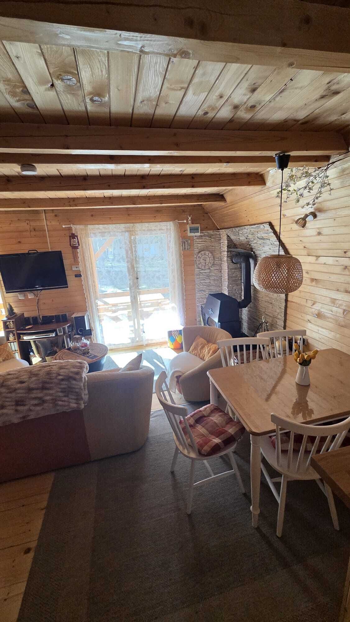 Un interior de cabaña acogedor con techo de madera, pared de piedra, chimenea, zona de comedor con mesa y sillas, y televisor montado en la pared.