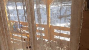 A room with a wooden floor, a window with a white lace curtain, and a view of a snowy outdoor area.