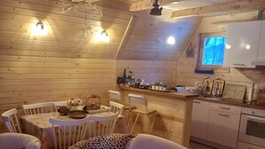 A cozy cabin interior with a wooden interior, a dining area with a table and chairs, a kitchen with cabinets and a window, and a sofa with cushions.