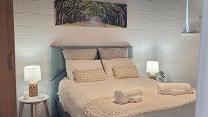 A bedroom with a bed, a ceiling fan, a wall-mounted painting, and a small side table with a lamp. - Cosy Reef View Apartment overlooking Sunset Coast, Fishing, Sand Dunes, Beaches. (Lancelin)