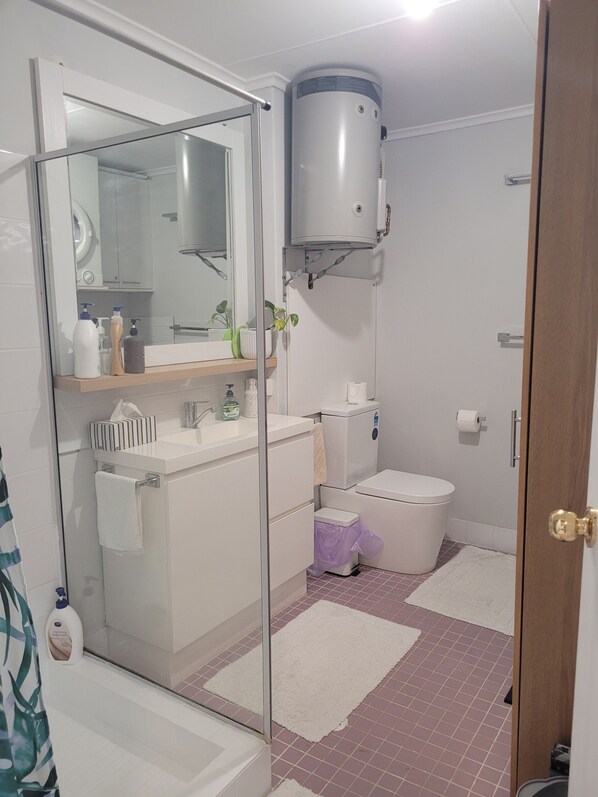 Un baño con cabina de ducha de vidrio, un inodoro, un lavabo con artículos de aseo y un suelo blanco con baldosas rosas.