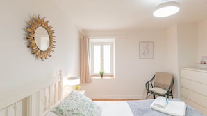 Un dormitorio con cama, silla, cómoda, espejo, ventana y una lámpara en el techo.