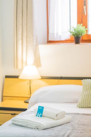 Un lit bien fait, avec une serviette pliée et une étiquette, une lampe de chevet, une tête de lit jaune et une fenêtre avec des rideaux.