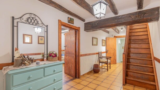 Un pasillo con escalera de madera, cómoda con cajones, espejo y puerta que da a otra habitación.