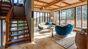 Un salón espacioso con una escalera de madera, ventanas amplias, un sofá, un sillón azul y una mesa de centro.