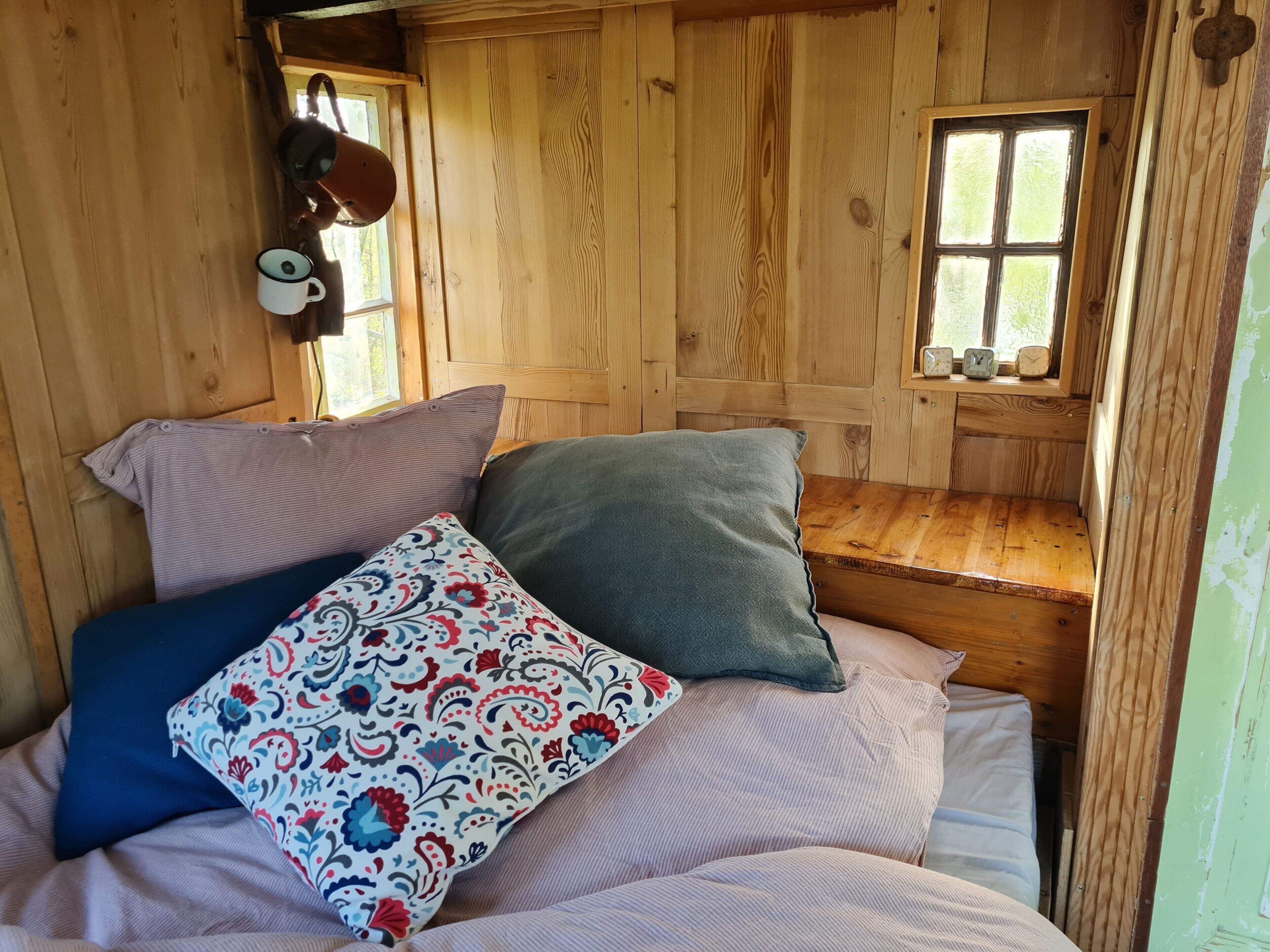 Ein Interieur einer Holzhütte mit einem Bett, Kissen, einem Fenster und einem kleinen Tisch mit einer Uhr.