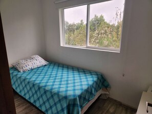 Une petite chambre avec un lit simple, une fenêtre, un lit recouvert d’une couverture à carreaux bleus et une table de chevet blanche.