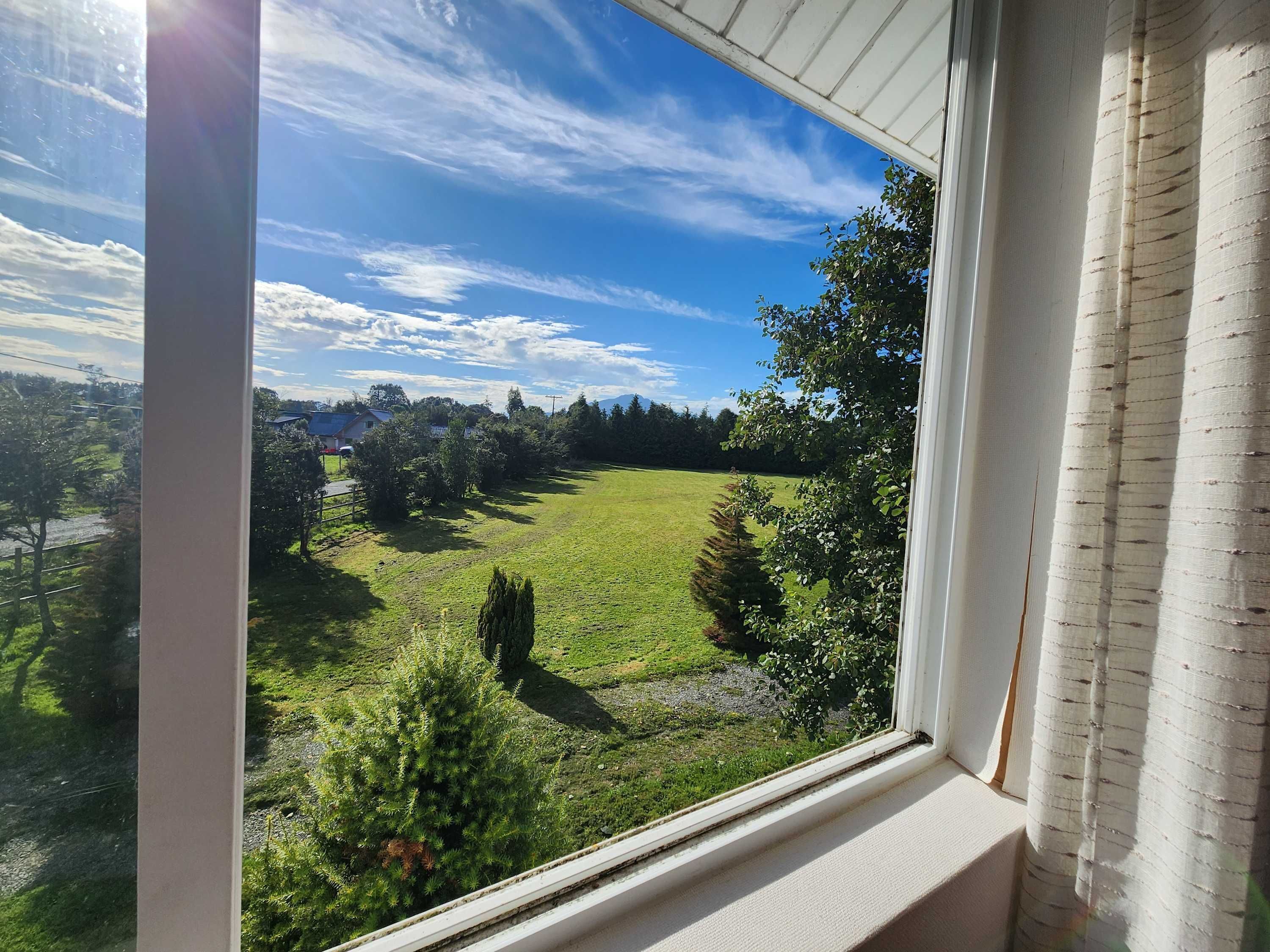 Una ventana con vistas a un campo cubierto de hierba, árboles y una casa a lo lejos.