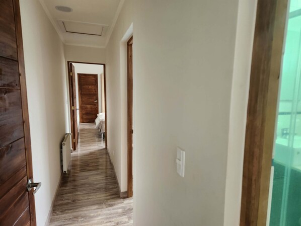 Un couloir étroit, doté d’un plancher en bois et de portes, qui mène à une pièce contenant un lit et une chaise.