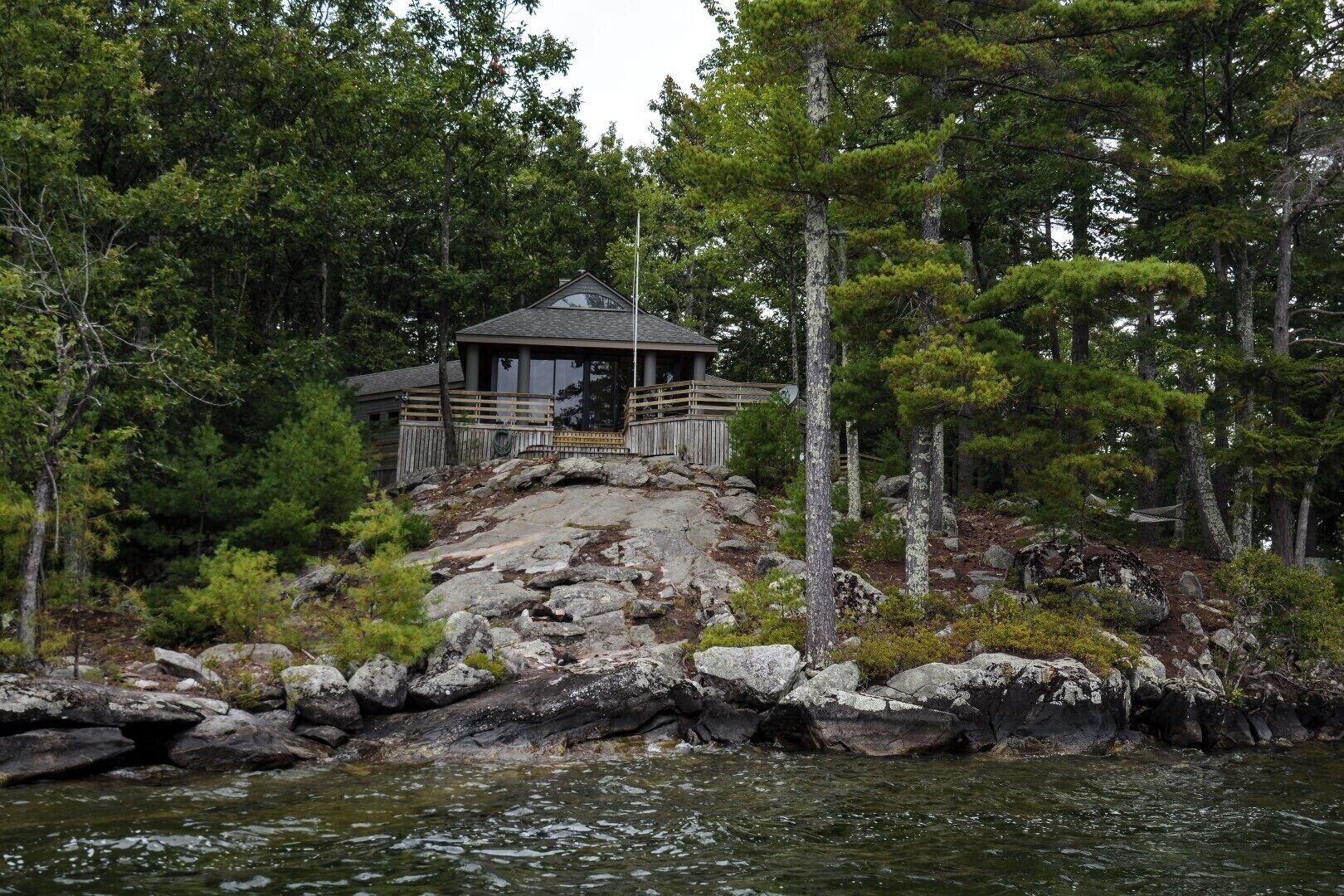 Eine Holzhütte mit einer Holzterrasse, umgeben von Bäumen und Felsen, mit Blick auf ein Gewässer.