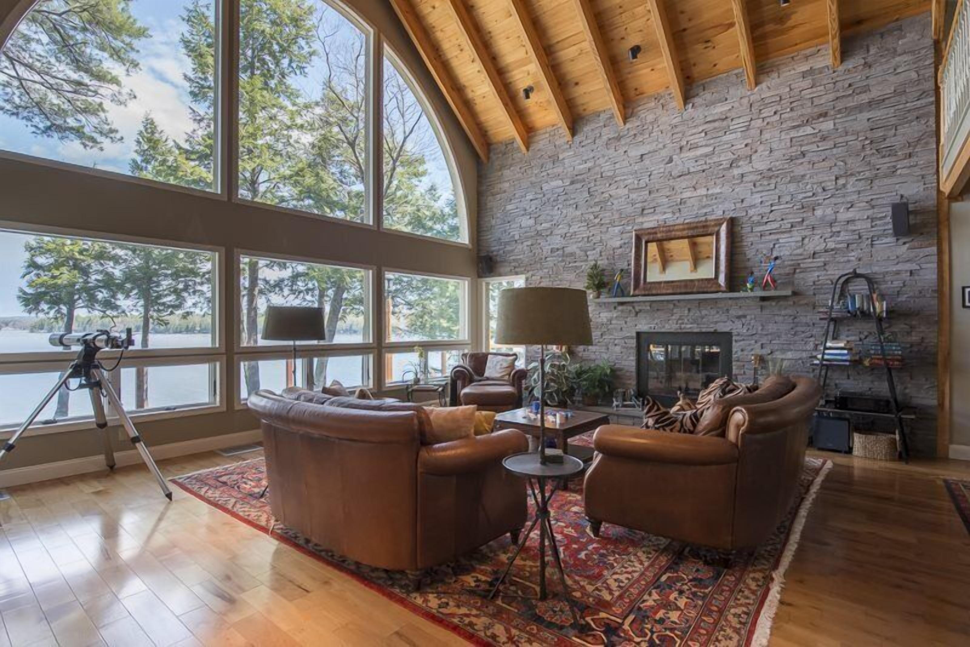 A spacious room with a stone wall, leather sofas, a fireplace, and large windows overlooking a body of water.