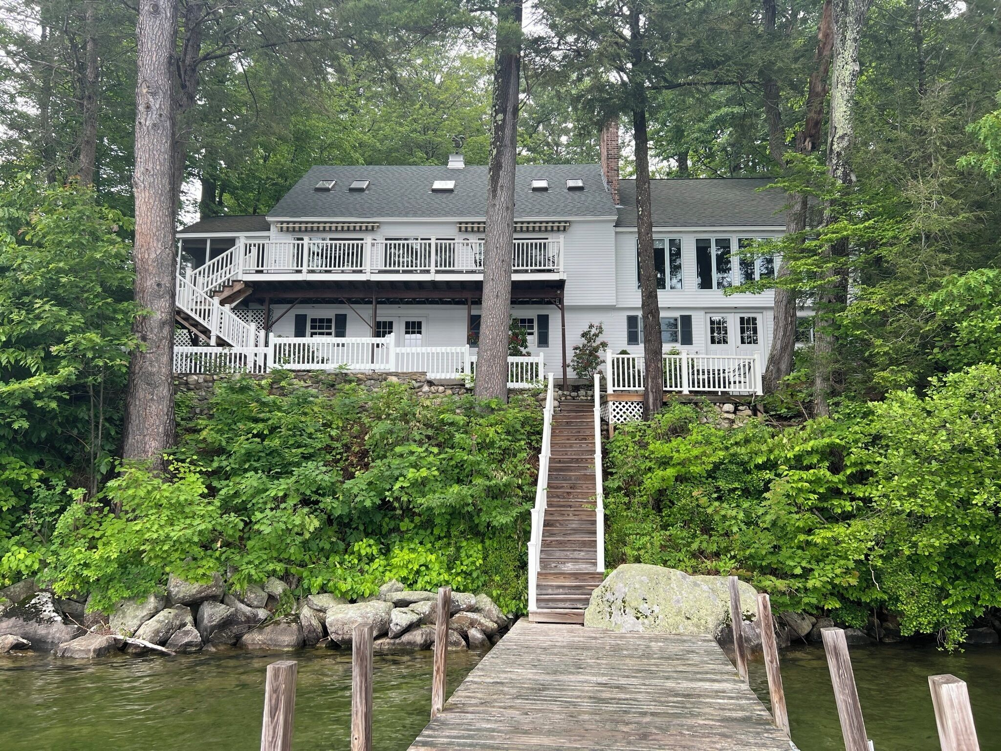 A two-story house with a wooden dock leading to a stone pier, surrounded by dense greenery and trees.
