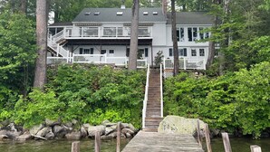 Une maison à deux étages avec une jetée en bois menant à un quai en pierre, le tout entouré d’une végétation luxuriante et d’arbres.
