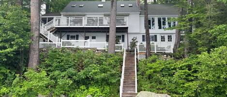 A two-story house with a wooden dock leading to a stone pier, surrounded by dense greenery and trees.